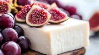Fresh figs and red grapes arranged with creamy white cheese on wooden board, macro food photography with soft natural lighting and shallow depth of field.