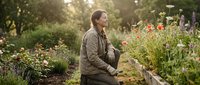 Young Caucasian woman gardener in brown jacket tending to colorful flower garden bed with blooming plants during golden hour sunlight for organic farming content.