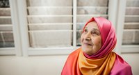 Elderly woman wearing pink and yellow hijab smiling indoors near window, representing senior diversity and cultural fashion for lifestyle and portrait projects.