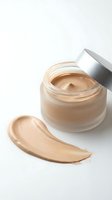 Open glass jar of beige foundation cream with silver cap and makeup swatch on white background for cosmetic product photography and beauty advertising.