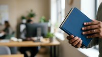 Closeup of hands holding closed blue book in modern office workspace with blurred desk and plants, suitable for education, learning, business training and professional development concepts.