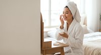 Young Asian woman in white bathrobe and towel applying skincare cream while looking in bedroom mirror during morning beauty routine.