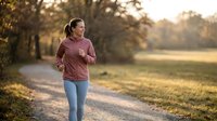 Young woman jogging on rural path during golden hour sunset wearing pink hoodie and jeans for fitness and healthy lifestyle outdoor exercise routine.
