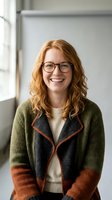 Smiling young Caucasian woman with curly red hair wearing glasses and colorful knit cardigan in modern office workspace for business and professional concepts.