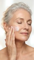 Mature woman with gray hair applying white moisturizer cream to face with closed eyes showing anti-aging skincare routine for healthy glowing skin.