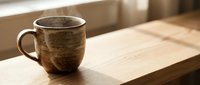 Rustic ceramic mug with textured glaze sitting on wooden table in natural sunlight, suitable for beverage, lifestyle, and home decor projects.