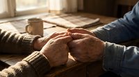 Elderly couple holding hands across table in warm sunlight, showing love, care and companionship in golden hour lighting with coffee and newspaper nearby.