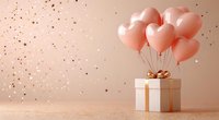 Pink heart-shaped balloons tied to white gift box with golden bow on beige background with glitter confetti, romantic celebration setup for Valentine's day or anniversary.