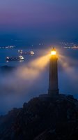 Lighthouse beacon shining through fog at dusk with illuminated ships in harbor creating atmospheric maritime scene with purple and blue twilight sky.