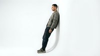 Young man with olive skin tone wearing casual olive green jacket and dark pants leaning against white curved wall. Isolated on white background for design use.