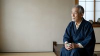 Senior Asian man in traditional dark blue kimono sitting indoors with contemplative expression, suitable for cultural, lifestyle, wellness, and editorial content.