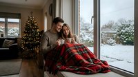Young couple wrapped in red plaid blanket enjoying hot drinks by window during winter holidays with Christmas tree and snowy landscape outside.
