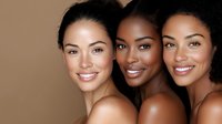 Three diverse women with natural makeup and glowing skin smiling confidently against neutral background, showcasing beauty, diversity and inclusivity in cosmetics industry.