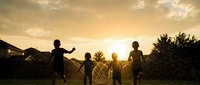 Four children playing together in backyard sprinklers during golden hour sunset, creating joyful summer memories with silhouettes against warm evening light.