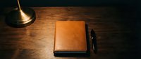 Leather-bound journal notebook with pen on wooden desk surface, illuminated by warm desk lamp light for writing, planning, and business organization.