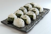 Traditional Japanese onigiri rice balls with nori seaweed and black sesame seeds arranged on dark stone plate against white background. Fresh Asian cuisine.