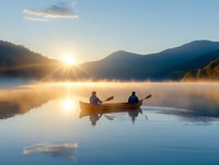 Two people paddling canoe on misty mountain lake at sunrise, silhouettes reflected in calm water with sunburst and fog creating atmospheric outdoor scene.