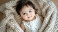 Adorable Asian baby girl in polka dot dress smiling while wrapped in cozy knit blanket, capturing pure joy and innocence in natural light setting.