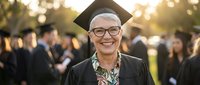 Happy senior Caucasian woman graduate in cap and gown smiling at outdoor ceremony with golden sunset light and blurred crowd background for education achievement.