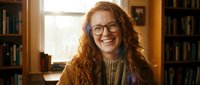 Happy young Caucasian woman with curly red hair and glasses smiling warmly in cozy library setting with books and natural window light for lifestyle content.