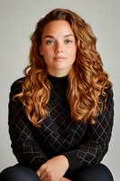 Young Caucasian woman with long wavy copper hair wearing black quilted sweater poses confidently against light background. Natural makeup and serene expression.