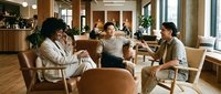 Diverse business team having casual meeting in modern office lounge with wooden furniture and natural lighting for collaborative workspace discussions.