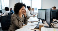 Stressed African American businesswoman with headache at office desk with computer monitors during busy workday.