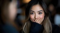 Young Asian woman covering mouth with black knitted sweater sleeve showing surprise or shyness with warm bokeh background for lifestyle content.