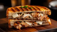 Grilled panini sandwich with melted cheese and roasted meat on wooden cutting board. Gourmet comfort food for restaurant menu and culinary marketing materials.