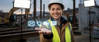 Male construction worker in safety helmet and yellow reflective vest holding digital communication icons above his palm at industrial building site.