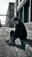 Young woman of mixed ethnicity sitting alone on urban street tying shoelaces, wearing black jacket and pants. Industrial warehouse buildings and metal bridge in background. Monochrome street photography.