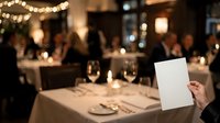 Waiter holding blank menu card at elegant dinner event with guests and warm string lights in background.