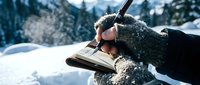 Gloved hand holding open book in snowy winter landscape with frosted trees, reading outdoors in cold weather conditions.