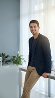 Confident young Caucasian businessman in casual blazer smiling while leaning on desk in modern bright office workspace for corporate marketing materials.