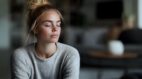 Young Caucasian woman with closed eyes and blonde hair in gray sweater sitting peacefully at home. Natural window light creates soft atmospheric mood for mindfulness concept.