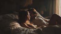 Mature couple relaxing together in bed reading book in warm morning sunlight streaming through window creating intimate cozy bedroom atmosphere.