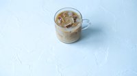 Iced latte in a clear glass mug on a white textured surface, with creamy milk swirling through cold brew coffee and ice cubes.