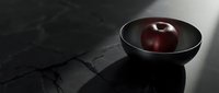 Dark red apple in black bowl on marble surface with dramatic lighting and shadow.