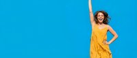Happy young woman in bright yellow dress celebrating with raised arm against vibrant blue sky background expressing joy and freedom outdoors.