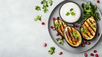 Grilled eggplant slices garnished with fresh herbs, pomegranate seeds and creamy yogurt sauce served on gray plate. Mediterranean cuisine background with copy space.