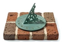 Vintage green patina sundial mounted on isolated red brick base with white mortar joints, casting shadow. Decorative garden ornament for outdoor landscaping, home decor, and architectural design projects.