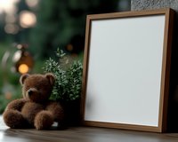 Brown teddy bear sitting next to wooden frame mockup with white blank space and green plant on blurred bokeh background, cozy home decoration setup.
