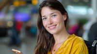 Young Caucasian woman in yellow blouse smiling warmly at camera with blurred urban background, natural makeup and long dark hair, professional headshot style portrait.