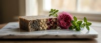 Handmade soap bar with oatmeal and dried botanicals beside fresh pink rose and green herbs on marble surface for natural skincare and wellness product marketing.