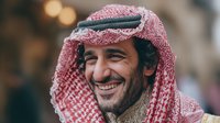 Smiling Middle Eastern man wearing traditional red and white keffiyeh headdress with black agal cord, showcasing authentic cultural attire against blurred urban background.