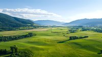 Rolling green hills and meadows stretch across valley between mountain ranges, sunlit agricultural landscape with scattered forests under blue summer sky.