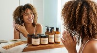 Young African American woman with curly hair applying skincare products in modern bathroom mirror reflection for daily beauty routine and self-care.