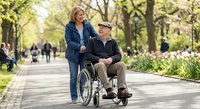 Caring blonde woman pushing elderly man in wheelchair through sunny park pathway with spring flowers and trees for healthcare assistance concept.