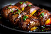 Grilled bacon-wrapped meat rolls stuffed with yellow bell peppers and garnished with fresh herbs on black plate, close up view with selective focus.