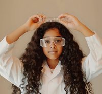 Young girl with curly hair wearing safety goggles and lab coat, adjusting protective eyewear during science experiment against neutral background.